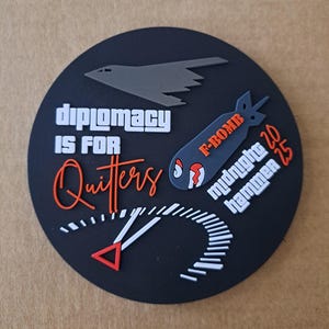 May include: A circular black patch featuring a grey stealth bomber silhouette, a grey bomb graphic labelled F-BOMB, and white text reading DIPLOMACY IS FOR Quitters, midnight hammer 20 25, alongside a white and red gauge graphic with a triangular needle.