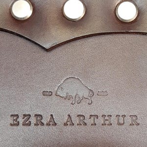 Puede incluir: Primer plano de una correa de cuero marrón con remaches plateados y una hebilla plateada. La correa presenta el texto en relieve "EZRA ARTHUR" debajo de un logotipo de bisonte. El cuero tiene una apariencia rica y texturizada.