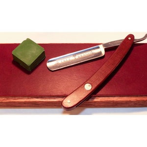 Puede incluir: Una navaja de afeitar con mango burdeos y hoja plateada, sobre un bloque de madera con superficie de cuero burdeos. También hay una piedra de afilar verde. La navaja está marcada "TREE BRAND".