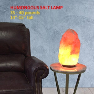 May include: A large, orange and yellow Himalayan salt lamp, weighing 35-40 pounds and standing 14-15 inches tall, sits on a gold metal stand with a white marble top. The lamp is in front of a brown leather armchair.