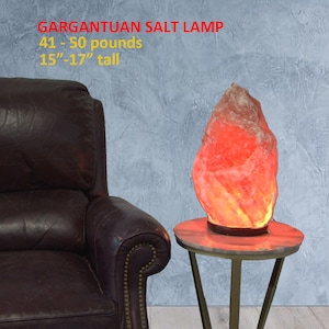 May include: A large, irregularly shaped Himalayan salt lamp, weighing 41-50 pounds and standing 15-17 inches tall. The lamp is lit and glowing orange, casting a warm light. It is sitting on a small, round, gold-colored table with three legs.