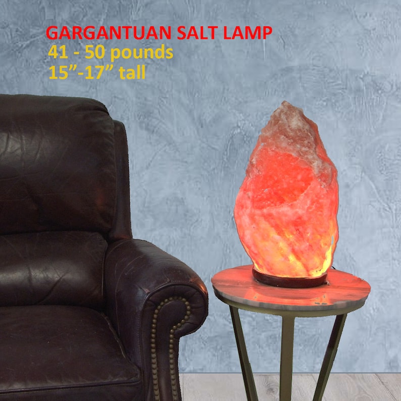 May include: A large, irregularly shaped Himalayan salt lamp, weighing 41-50 pounds and standing 15-17 inches tall. The lamp is lit and glowing orange, casting a warm light. It is sitting on a small, round, gold-colored table with three legs.