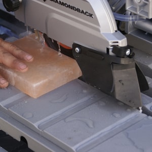 May include: A close-up of a gray wet saw with a pink salt block being cut. The saw is labeled "DIAMONDBACK".