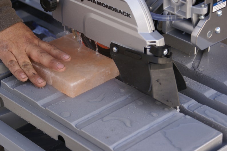May include: A close-up of a gray wet saw with a pink salt block being cut. The saw is labeled "DIAMONDBACK".