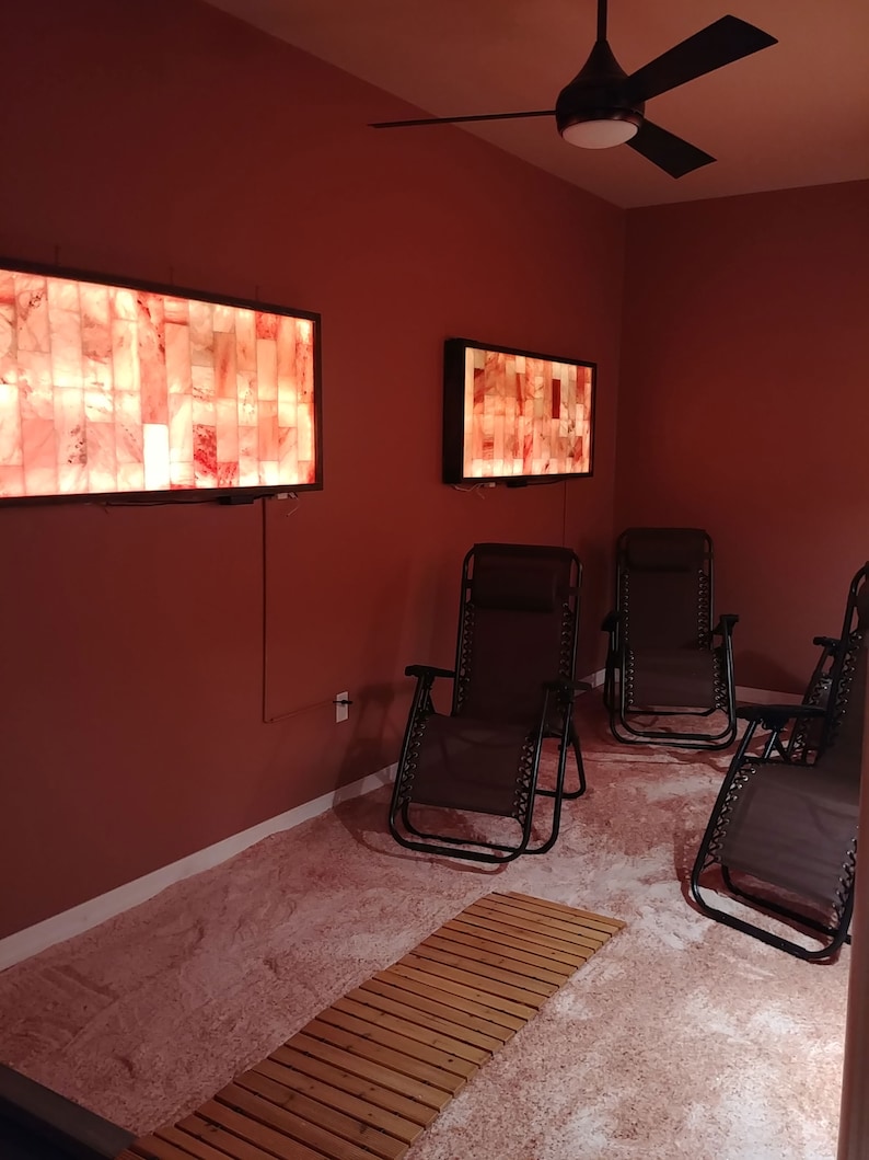 May include: A salt therapy room with a pink Himalayan salt floor, four black reclining chairs, and three salt lamps on the walls.