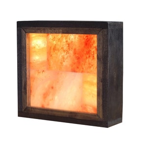 May include: A square Himalayan salt lamp with a wooden frame. The salt is a warm orange color and is illuminated from within, creating a soft glow.
