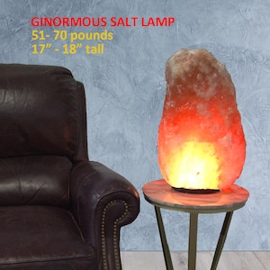 May include: A large, orange-hued Himalayan salt lamp, weighing 51-70 pounds and standing 17-18 inches tall, sits on a gold metal stand with a white marble top. The lamp is lit, casting a warm glow. The lamp is in front of a brown leather armchair.