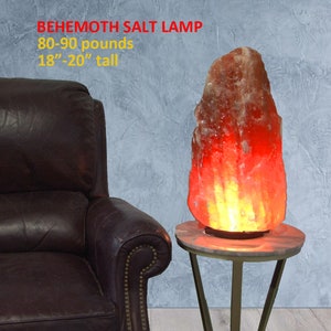 May include: A large, reddish-orange Himalayan salt lamp, weighing 80-90 pounds and standing 18-20 inches tall, is lit and glowing on a gold metal stand with a round white top. The lamp is in front of a brown leather armchair.