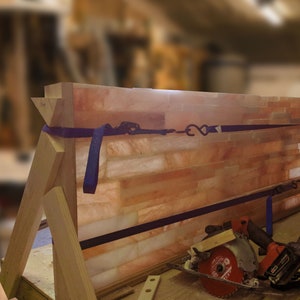 May include: A wooden frame holds a large panel of Himalayan salt bricks. The salt bricks are a light orange color and have a rough texture. A blue strap is wrapped around the frame and secured with a metal hook. A circular saw and a power drill are lying on a wooden table in front of the frame.