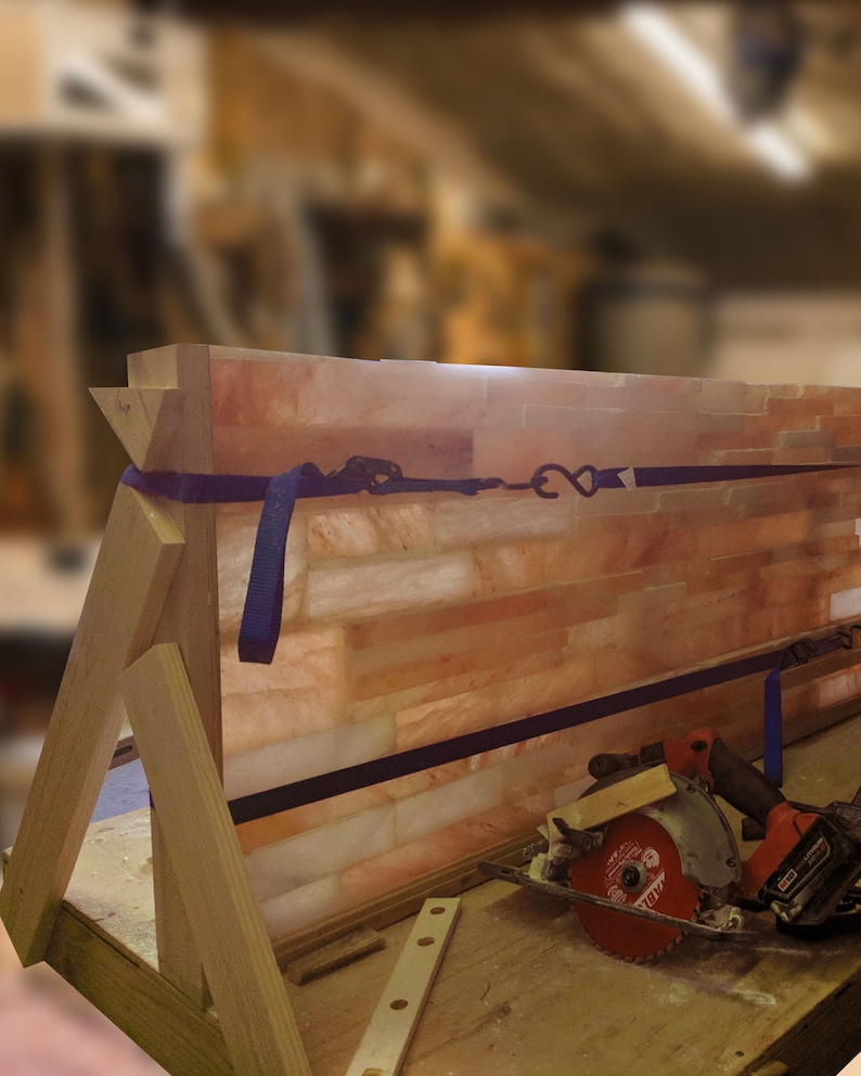 May include: A wooden frame holds a large panel of Himalayan salt bricks. The salt bricks are a light orange color and have a rough texture. A blue strap is wrapped around the frame and secured with a metal hook. A circular saw and a power drill are lying on a wooden table in front of the frame.