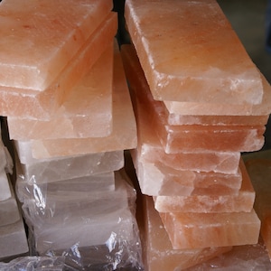 May include: A stack of pink Himalayan salt slabs, some are smooth and others have a rough texture. The slabs are stacked in a pyramid shape.