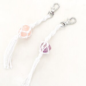 May include: Two macrame keychains with silver clasps. One features a pink rose quartz crystal, the other a purple amethyst crystal. The crystals are held in a white macrame net, with a white tassel at the end. Ideal for keys or bags.