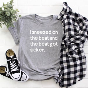 May include: A grey t-shirt with the text "I sneezed on the beat and the beat got sicker." in white lettering. The shirt is laid flat on a white brick background with a pair of black trainers and a black and white checked shirt.