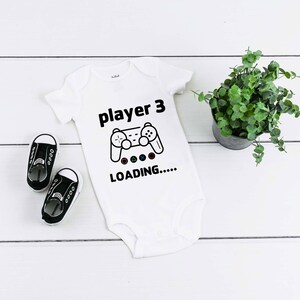 May include: A white short-sleeved baby bodysuit featuring the black text player 3, a black line-art illustration of a video game controller, and the text LOADING..... with small colored geometric shapes underneath the controller graphic.