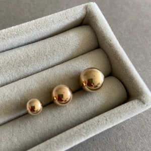 May include: Three gold-colored ball earrings of different sizes displayed in a gray velvet box.