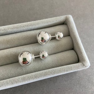May include: A pair of silver ball earrings with a post and a larger ball on the front. The earrings are displayed in a gray velvet box.
