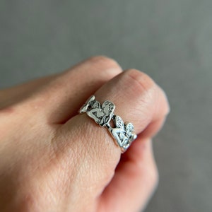 K&ouml;nnte beinhalten: Ein silberner Ring mit einem Design aus mehreren Cherubim oder Engeln, die miteinander verbunden sind. Der Ring wird an einem Finger getragen.