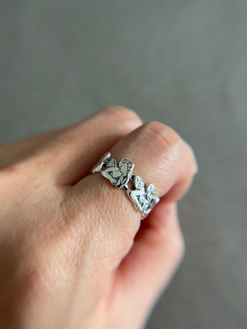 K&ouml;nnte beinhalten: Ein silberner Ring mit einem Design aus mehreren Cherubim oder Engeln, die miteinander verbunden sind. Der Ring wird an einem Finger getragen.