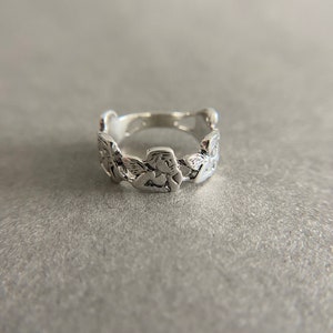 K&ouml;nnte beinhalten: Ein silberner Ring mit einem einzigartigen Design, das aus einer Reihe von verbundenen, strukturierten, blattartigen Formen besteht.