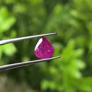 May include: A vibrant, teardrop-shaped magenta gemstone held by a pair of silver-toned metal tweezers. The stone features a faceted surface that catches the light, displaying a deep pink hue with internal textures visible through the translucent material.