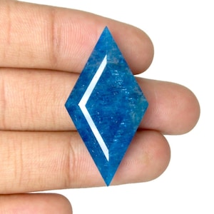 Puede incluir: Una gema en forma de diamante de un vibrante color azul. La gema presenta una incrustación blanca y angular que acentúa su forma geométrica. La piedra se sostiene en una mano, mostrando su tamaño y color sobre un fondo neutro.