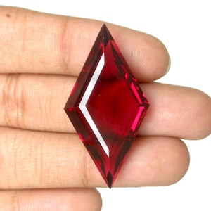 Puede incluir: Una gema facetada en forma de diamante, de color rojo intenso. La gema se sostiene en una mano sobre un fondo blanco. La gema tiene ángulos agudos y una superficie pulida que refleja la luz.