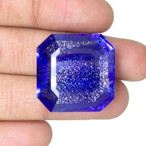 Könnte beinhalten: Ein großer, quadratisch geschliffener Edelstein mit tiefblauer Farbe und einem glitzernden Inneren. Der Stein wird in einer Hand vor weißem Hintergrund gehalten, um seine Farbe und Klarheit hervorzuheben. Die Facetten reflektieren das Licht und erzeugen ein brillantes Aussehen.
