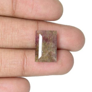 Può includere: Una gemma rettangolare con una miscela di colori, tra cui rosa, marrone e bianco. La gemma ha un taglio sfaccettato con un design geometrico bianco. La pietra è tenuta in una mano.