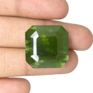 以下が含まれることがあります： 八角形のファセット加工が施されたエメラルドグリーンの宝石。白い背景を背景に指で宝石を持ち、その色と透明度を強調しています。石には内部の静脈があり、明るく反射する表面があります。