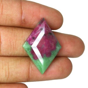 Puede incluir: Una piedra preciosa de rubí zoisita en forma de cometa con una superficie facetada, que presenta una mezcla vibrante de colores magenta rosado intenso y verde bosque, con inclusiones naturales y un acabado pulido que refleja la luz.