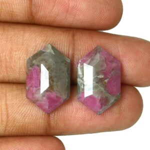 May include: Two loose, hexagonal-cut gemstones featuring a mix of opaque grey and vibrant pink inclusions. These faceted stones have a flat top surface and a geometric shape, displaying a natural, mottled colour pattern throughout the crystalline structure.