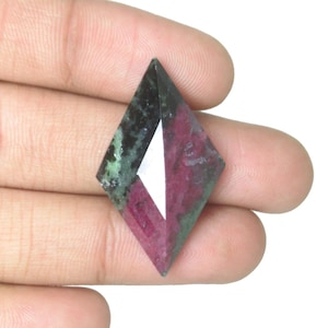 Puede incluir: Una gema facetada en forma de diamante, con un llamativo contraste de colores. Una mitad muestra un tono rojo intenso, mientras que la otra presenta una mezcla de verde y negro. La gema se sostiene en una mano.