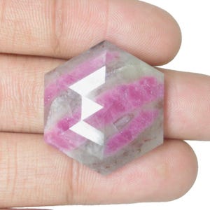Peut inclure: Une pierre précieuse hexagonale facettée, avec des nuances claires, grises et roses. La gemme présente un motif en zigzag blanc et des bandes roses. La pierre est tenue dans une main.