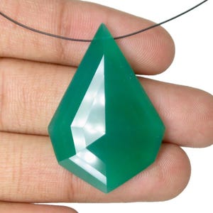 May include: A faceted, emerald-green gemstone pendant with a geometric shape. The pendant is suspended from a thin, dark cord. The gemstone is held in a hand, showcasing its polished surface and sharp angles. The pendant is a striking piece of jewellery.
