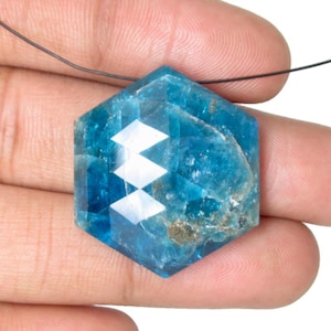 Puede incluir: Un colgante hexagonal de piedra preciosa azul facetada. La gema es de un color turquesa vibrante con un patrón geométrico de facetas blancas. Está ensartado en un fino cordón negro, lo que sugiere que es un collar.