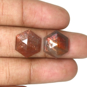 May include: Two hexagonal sunstone gemstones with a faceted surface. The stones exhibit a reddish-brown hue with internal metallic inclusions that create a shimmering effect. Each gemstone features a geometric six-sided cut with sharp edges.