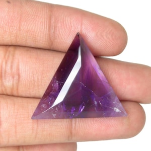 May include: A triangular amethyst gemstone with a polished surface. The gem is a deep purple color with lighter purple and white highlights. The facets of the stone catch the light, creating a sparkling effect. The gemstone is held in a hand.