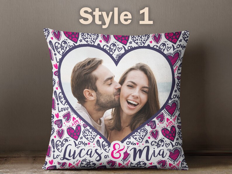 Puede incluir: Una almohada blanca con un marco de fotos en forma de coraz&oacute;n que presenta a una pareja. La almohada tiene un patr&oacute;n de corazones rosa y morado y las palabras "Love" y "Lucas & Mia".