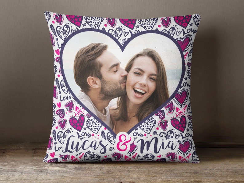 Puede incluir: Una almohada blanca con una foto en forma de coraz&oacute;n de una pareja bes&aacute;ndose. La almohada est&aacute; decorada con corazones rosas y morados y las palabras "Lucas & Mia".