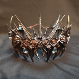 May include: A dark, ornate crown with a gothic design. The crown features sharp, pointed elements and small, round, copper-colored accents. The metal has a dark, weathered appearance, suggesting an antique or fantasy aesthetic. The crown is designed to be worn.