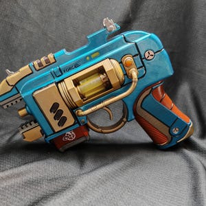 May include: A futuristic-looking toy gun with a teal and gold color scheme. The gun features intricate details, including a trigger, barrel, and various mechanical components. The word "FORCE" is visible on the side. The handle is brown.