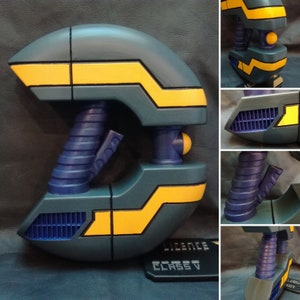 Puede incluir: Un dispositivo futurista y port&aacute;til en tonos de verde azulado, azul marino y amarillo. El dise&ntilde;o presenta formas geom&eacute;tricas, un asa acanalada y un peque&ntilde;o detalle esf&eacute;rico. El dispositivo tiene una estructura similar a una rejilla y el texto "LICENCE CLASS V" es visible.