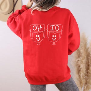 May include: Red sweatshirt with a white graphic of two smiling Ohio state outlines holding signs that say "OH" and "IO".