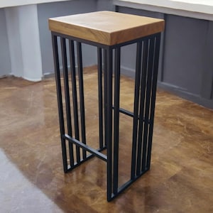 Op de afbeelding: Een hoge, vierkante barkruk met een lichtbruine houten zitting en een zwart metalen frame. Het frame heeft verticale staven en een horizontale staaf nabij de basis. De kruk is ontworpen voor een moderne uitstraling.