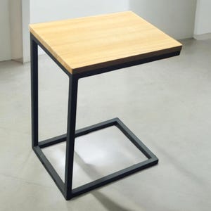 May include: A contemporary side table featuring a light wood surface and a black metal frame. The table has a square top and a C-shaped design, with a base that is 38 cm by 38 cm.