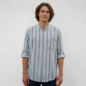 May include: A light blue and white striped shirt with a mandarin collar and a single chest pocket. The long sleeves are rolled up. The shirt is paired with dark blue jeans.