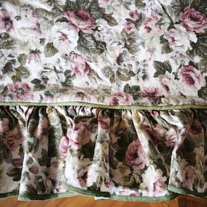 Puede incluir: Tela floral con un estampado de rosas en tonos rosa, blanco y verde. La tela tiene un borde con volantes y ribete verde. El diseño es de estilo vintage y romántico.