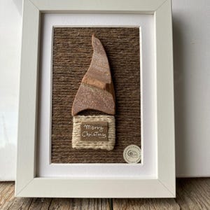 Op de afbeelding: Een ingelijst kunstwerk met een bruine en beige gnome gemaakt van natuurlijke materialen. De gnome draagt ​​een bruine hoed en heeft een witte baard. Het kunstwerk is op een bruine achtergrond en heeft de woorden "Prettige Kerst" erop geschreven.