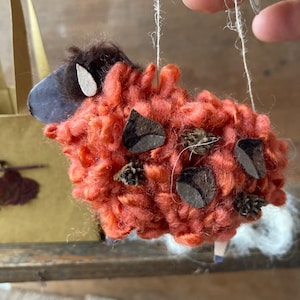 Può includere: Un ornamento di pecora in lana arancione fatto a mano con dettagli in feltro marrone e un orecchio in feltro bianco. La pecora è appesa a una corda.