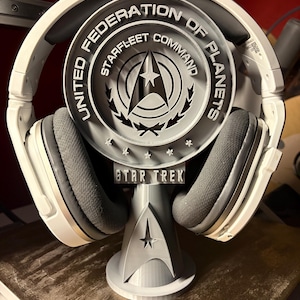 May include: A gray Star Trek-themed headphone stand with the words "United Federation of Planets Starfleet Command" and a Starfleet insignia. The stand holds white and gray headphones.